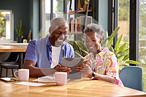 Smiling Senior Couple Sitting Around Table At Home Reviewing Finances Using Digital Tablet