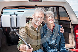 smiling senior couple sitting on car, embracing and taking selfie.