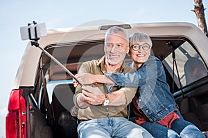 smiling senior couple sitting on car, embracing and taking selfie.