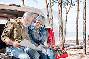 smiling senior couple sitting on car and holding map in sunny day.