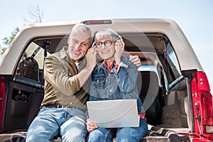 smiling senior couple of tourists with laptop listening music in headphones in car.