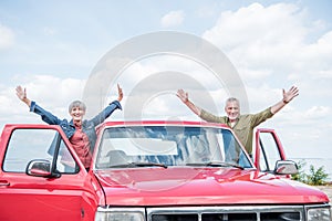 smiling senior couple of tourists standing with hands up near red car at beach.