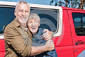 smiling senior couple of tourists standing near red car and embracing in sunny day.