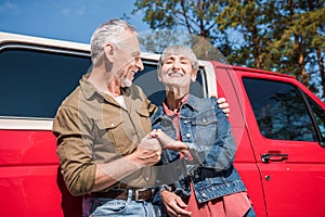 smiling senior couple of tourists standing near red car, holding hands and embracing in sunny day.