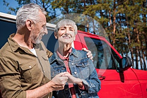 smiling senior couple of tourists standing near red car, holding hands and embracing in sunny day.