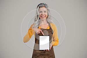 Smiling senior european woman gray hair framer in apron point finger at banner with free space