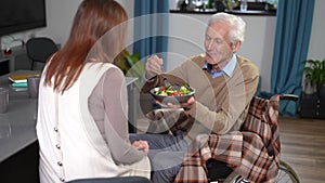 Smiling senior husband in wheelchair feeding wife with vegetable salad at home. Portrait of happy smiling Caucasian man