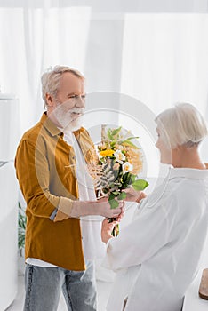 Smiling senior man giving bouquet to wife on blurred foreground in kitchen.