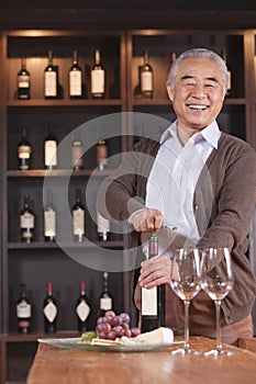 Smiling senior man opening wine bottle and looking at camera, shelf with wine in the background