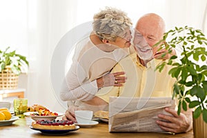 Smiling senior man reading newspaper and his wife kissing him