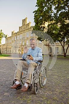 Smiling senior man, recovering male patient in wheelchair making a video call using smartphone while resting alone in