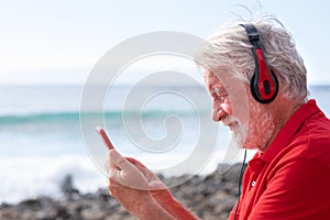 Smiling senior man with white hair and beard having video conversation on mobile phone. Sitting on the rock beach facing the sea