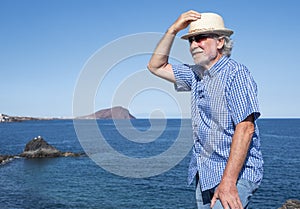 Smiling senior man white haired wearing a straw hat standing and enjoying vacation and freedom in seascape. Blue sea red mountain