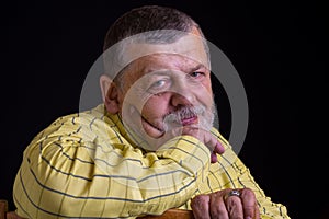 Smiling senior man in yellow shirt leant elbow on  a back of chair against dark background