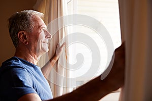 Smiling senior white man opening the curtains on a sunny morning, side view, close up