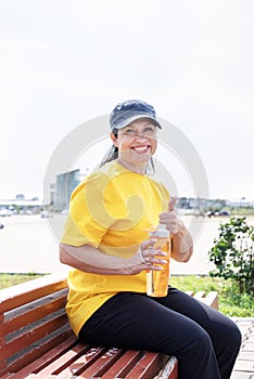 Smiling senior woman drinking water after workout outdoors on the sports ground showing thumbs up