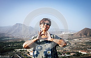 Smiling senior woman enjoying freedom standing on hill with mountain and city on background looking at camera making heart shape