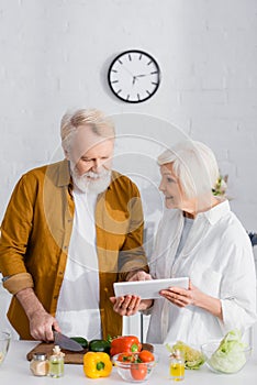 Smiling senior woman holding digital tablet near husband cooking in kitchen.