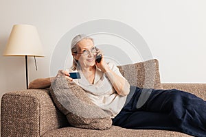 Smiling senior woman making a call via mobile phone and holding a credit card while lying on sofa