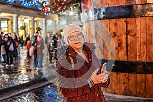 Smiling senior woman in purple jacket walks down the central city a rainy night street and uses her phone. Christmas