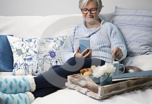 Smiling senior woman relax in sofa at home using smartphone and having a break with food and drink