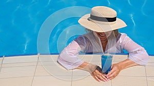 Smiling senior woman relaxing near the blue outdoor swimming pool with a blue cocktail. People are enjoying their summer