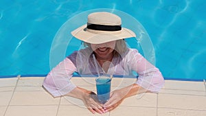 Smiling senior woman relaxing near the blue outdoor swimming pool with a blue cocktail. People are enjoying their summer