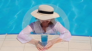 Smiling senior woman relaxing near the blue outdoor swimming pool with a blue cocktail. People are enjoying their summer