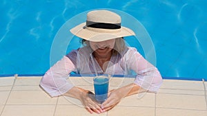 Smiling senior woman relaxing near the blue outdoor swimming pool with a blue cocktail. People are enjoying their summer
