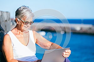 Smiling senior woman sitting outdoor at sea port using laptop computer. Relaxed elderly woman wearing sunglasses, sunset light