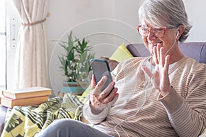 Smiling senior woman sitting on sofa at home in video call with mobile phone. Wearing earphones and eyeglasses