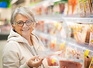 A smiling senior woman at the supermarket chooses which ham to buy. Many products on offer in the refrigerated counter