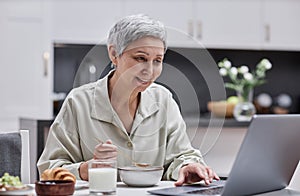 Smiling senior woman using laptop and working while enjoying breakfast at home