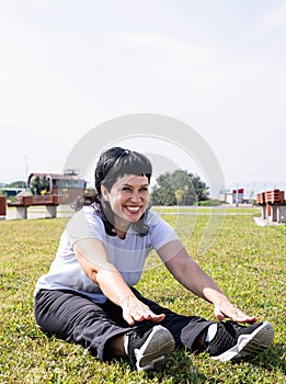 Smiling senior woman warming up stretching sitting on the grass in the park