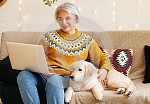 Smiling senior woman watch Christmas movie on laptop, relaxing on couch with golden retriever puppy