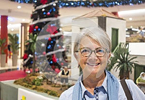 Smiling senior woman white hair and glasses standing in shopping mall with christmas tree on background - concept of holidays and