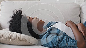 Smiling serene African young woman head hits pillow, stress-free millennial female student untroubled relaxing on couch