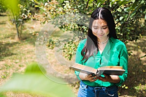Smiling serene young woman reading a book in the greenery of the field. World book day. Copy space