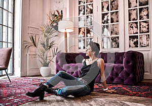 Smiling short haired brunette woman in black shirt, jeans and brutal shoes sits on the floor in the centre of room