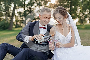 Smiling and shy groom and bride drinking and pouring bubbly champagne wine from bottle to glasses together, sit in park