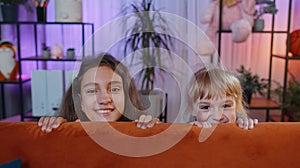 Smiling siblings children girls playing hide and seek peekaboo game trying to scare, prank at home