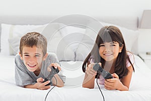 Smiling siblings lying on bed playing video games together