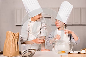 smiling sister and brother in chef hats and aprons whisking eggs in bowl at table