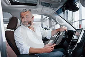 Smiling while sitting in a brand new car. Bearded senior man in white shirt holds mobile phone