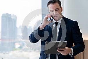 Smiling skilled businessman talking phone while looking at data. Ornamented.