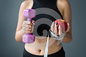 woman in holding apple and measuring tape and dumbbell standing  over gray background