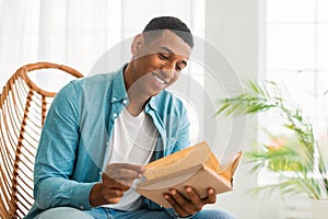 Smiling smart millennial african american guy reading book, sit on chair, enjoy comfort and free time photo