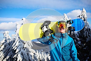 Smiling snowboarder posing carrying snowboard on shoulder at ski resort near forest before backcountry freeride and