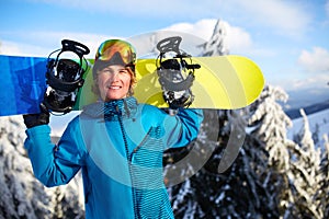 Smiling snowboarder posing carrying snowboard on shoulders at ski resort near forest before backcountry freeride and