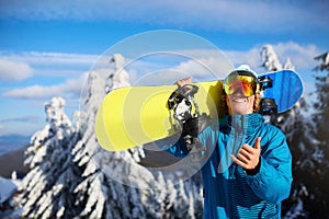 Smiling snowboarder posing carrying snowboard on shoulders at ski resort near forest before freeride session. Rider